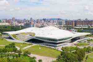 衛武營國家藝術文化中心場館外觀，白色波浪狀屋頂建築座落於草坡園區，後方為高雄市區天際線